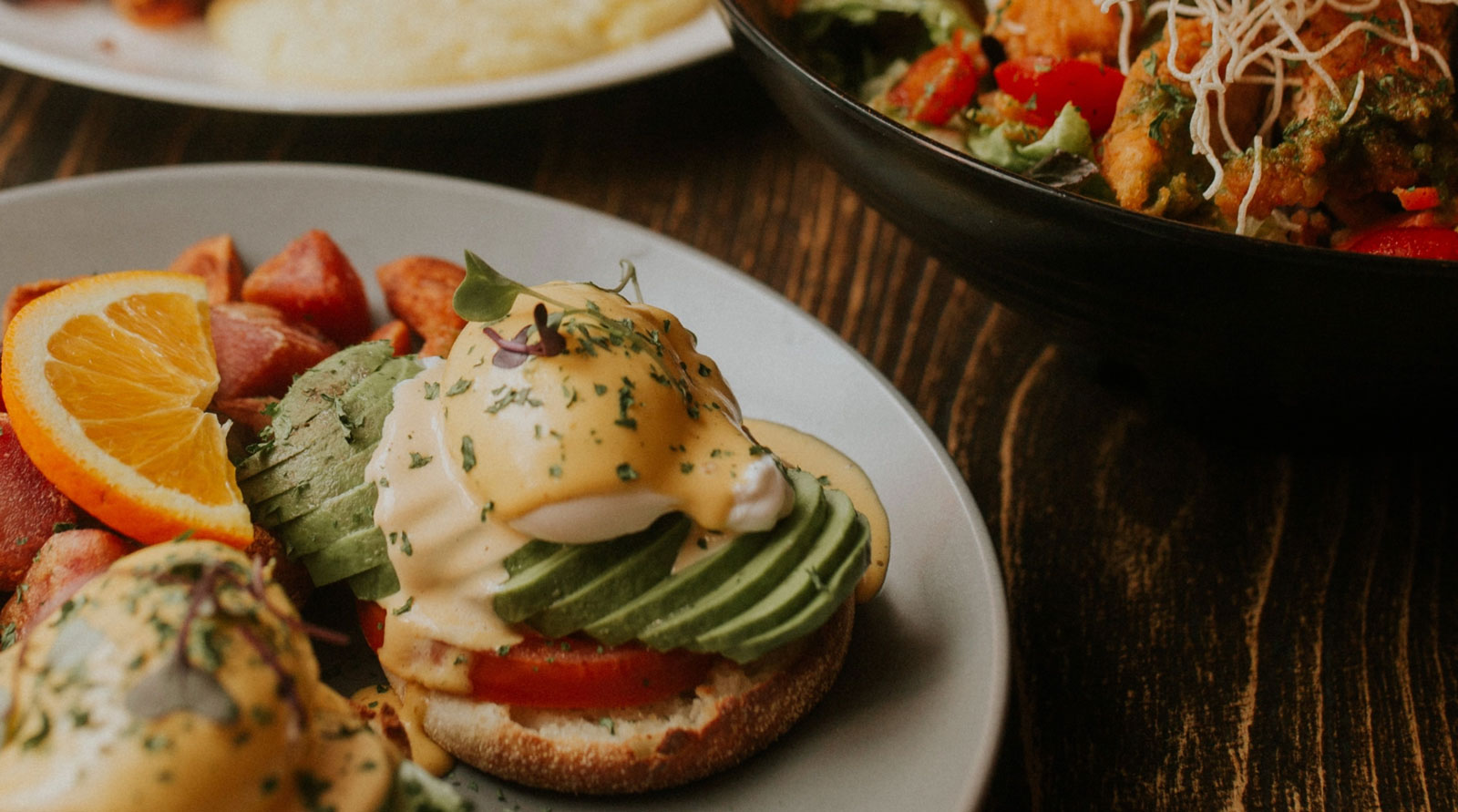 Pictured: a brunch plate filled with eggs benedict. From bottom to top: toasted crostini bread, fresh sliced tomato, generous slices of avocado, and a perfectly poached egg topped with Hollandaise sauce, fresh herbs, and microgreens. Also on the plate are a slice of fresh orange and a pile of perfectly browned golden home fried potatoes.