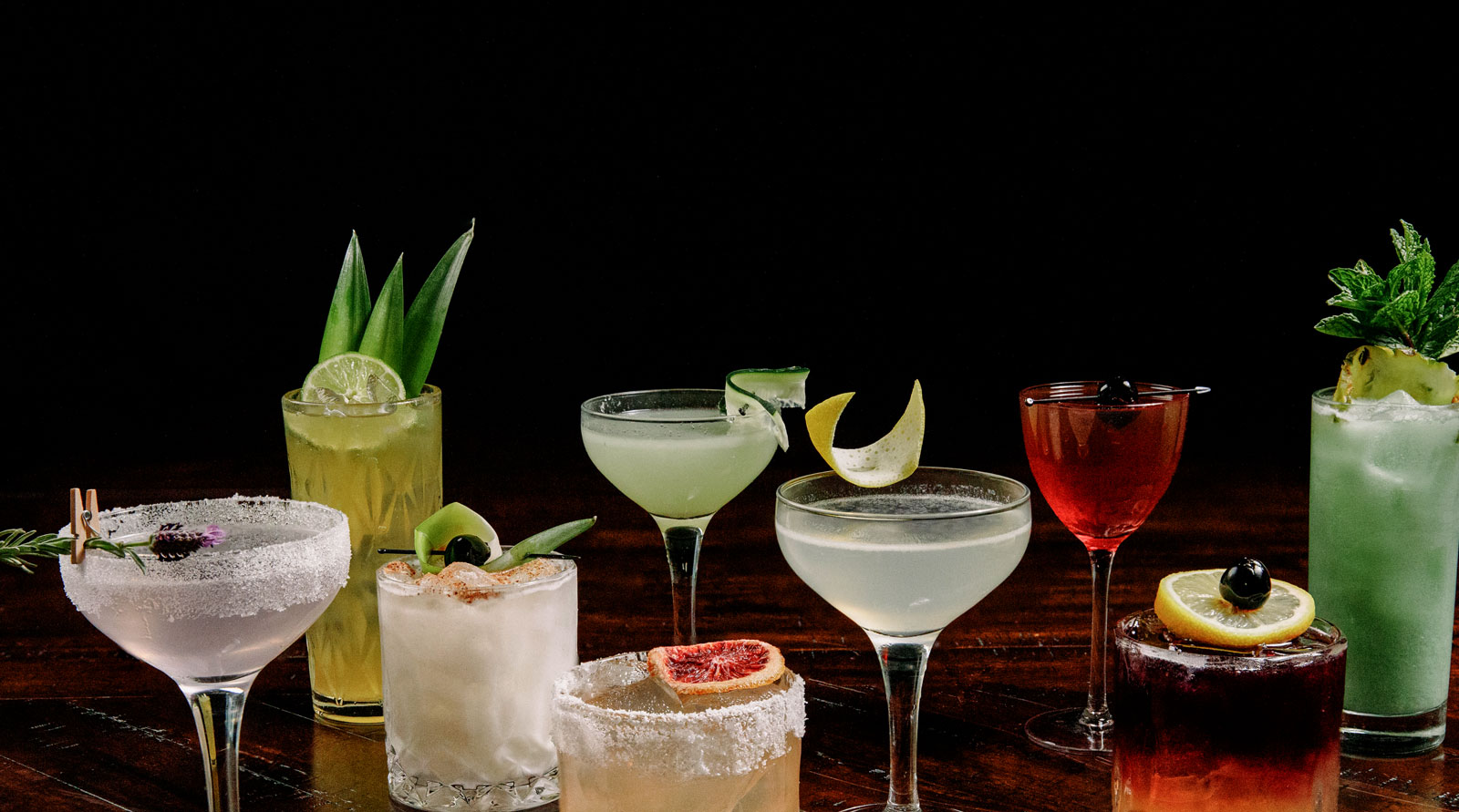 Pictured: an array of seasonal cocktails including margaritas, mojitos, mules, and more. Some of the drinks feature sugared rims. Other garnishes include sprigs of lavender, slices of lime and blood orange, delicate curls of citrus peel, and fresh mint.