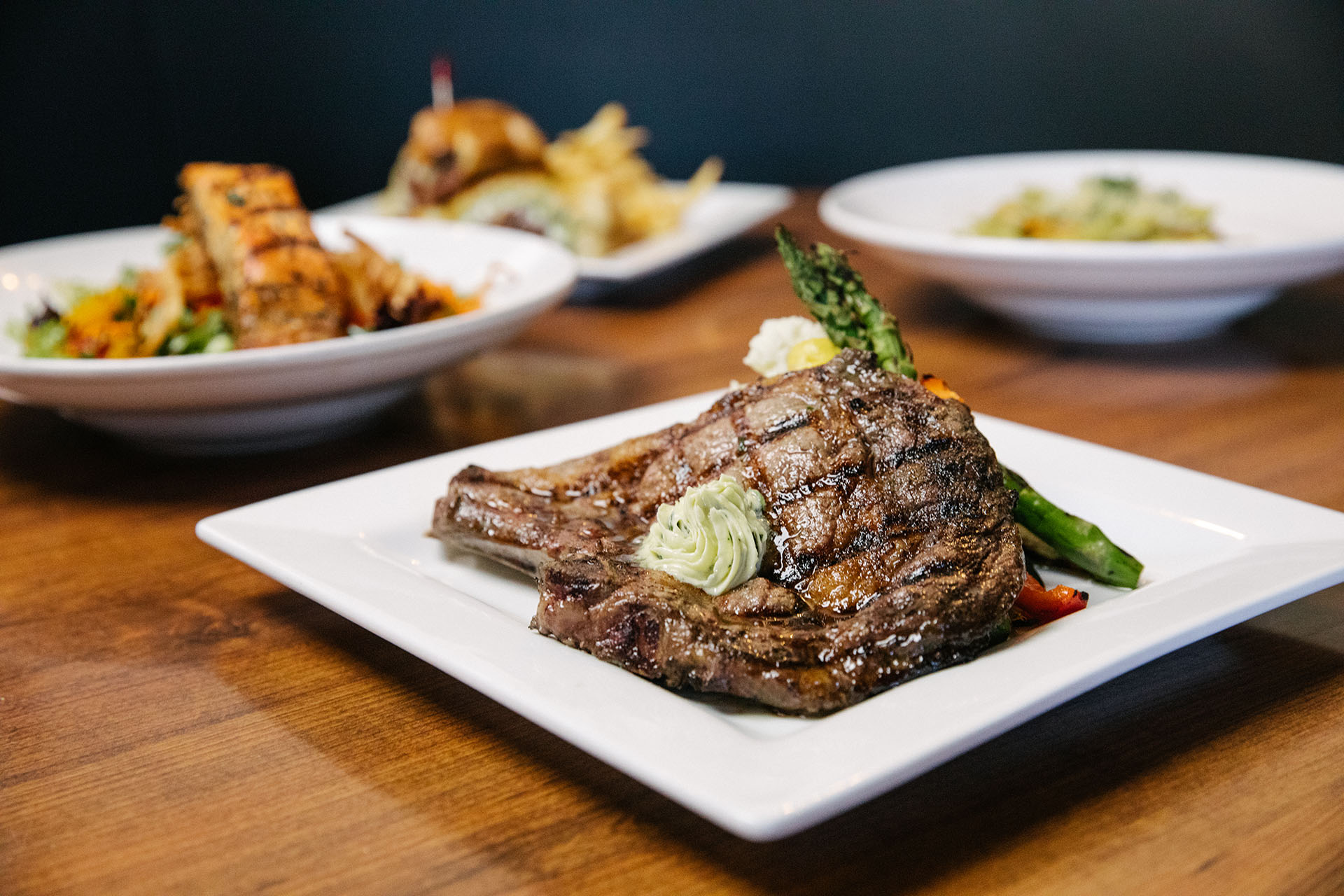 Pictured: our delicious flat iron steak entree with Chimichurri, Fried Spring Onion, Shallot, Mashed Potatoes, and Charred Vegetables. The steak is perfectly browned and glistening and takes up almost the entire plate. Visit Piazza del Pane for Fresno's best Italian dining and more.